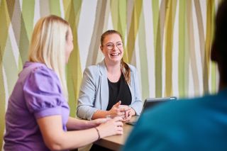 Een lachende, vrolijke woonassistent in een huiskamer met groen behang in gesprek met twee andere zorgmedewerkers.